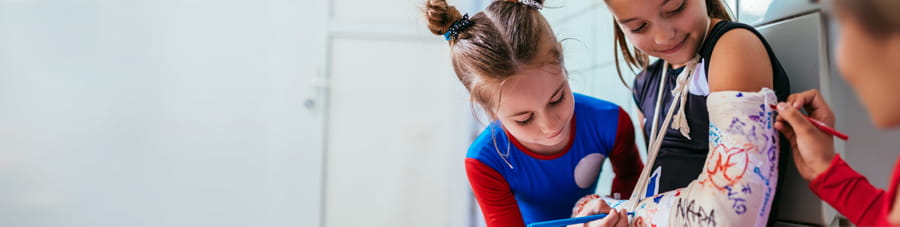 Two young girls sign another girl's arm cast.