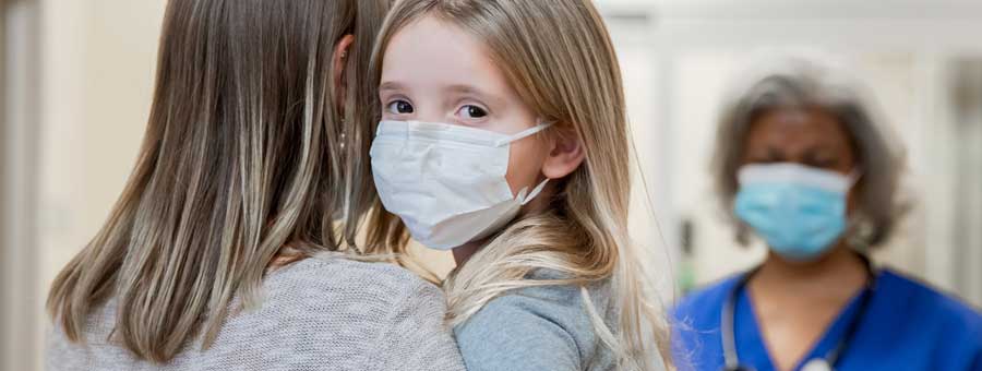 Young child in mask is held by an adult while a health care worker in a mask looks on