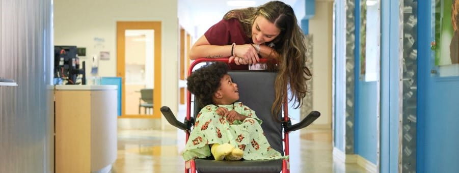 Health care worker pushes child patient down hospital hallway in wheelchair.